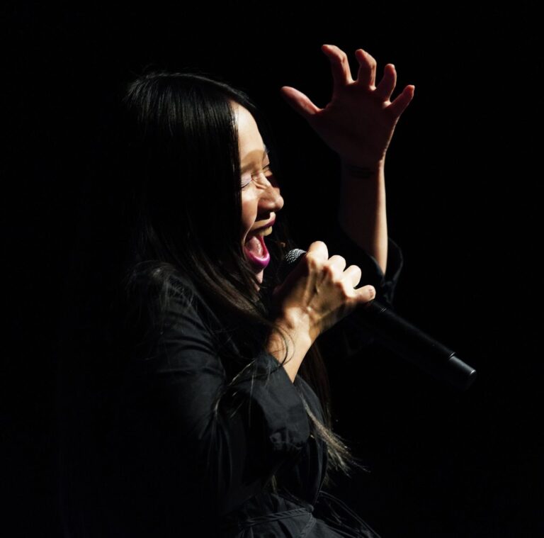 A woman with long dark hair performs on a darkened stage, lit dramatically from one side against a black background. She holds a microphone close to her open mouth, her expression intense and expressive. Her other hand is raised above her head, fingers spread wide, conveying passion and emotional force. She wears a black outfit and bright pink lipstick. The photograph captures a moment of powerful live performance — singing, spoken word, or theatre.